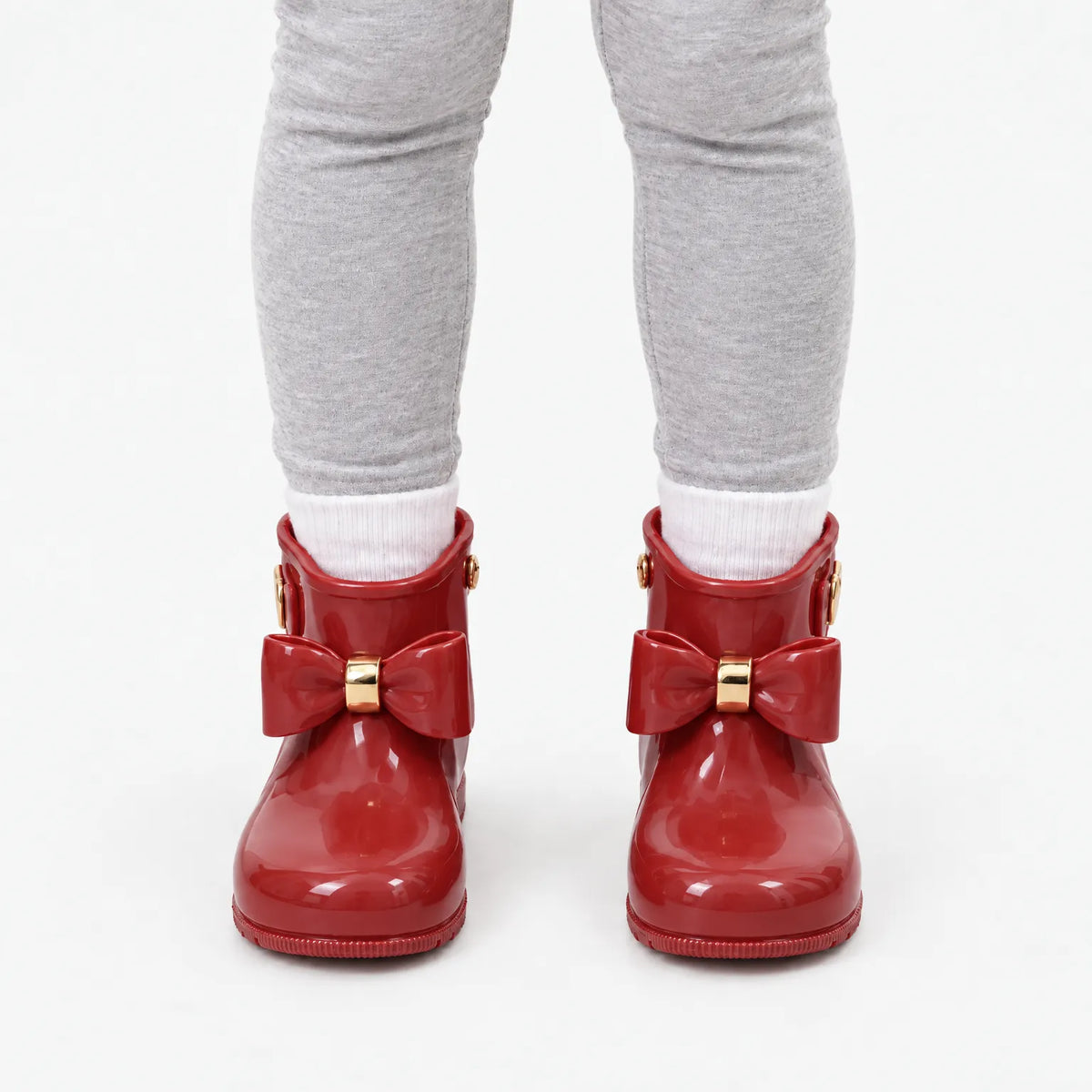 Front view of a child wearing glossy red bow wellies with white socks and grey leggings. 10