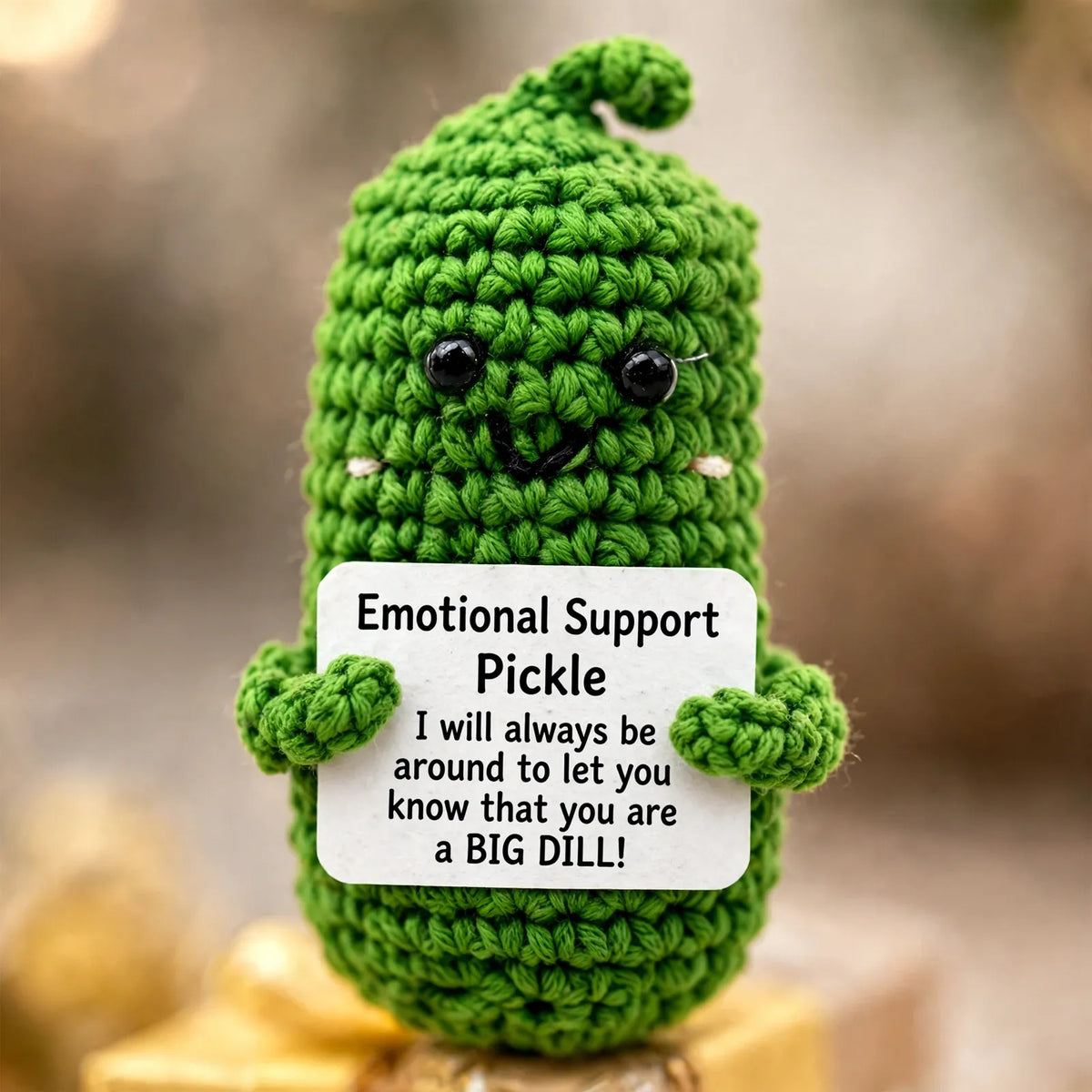 Close-up of the green crocheted pickle character with black bead eyes, holding a white message card 3
