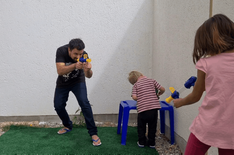 A man and a girl point toy guns outdoors while a toddler leans over a blue stool 10