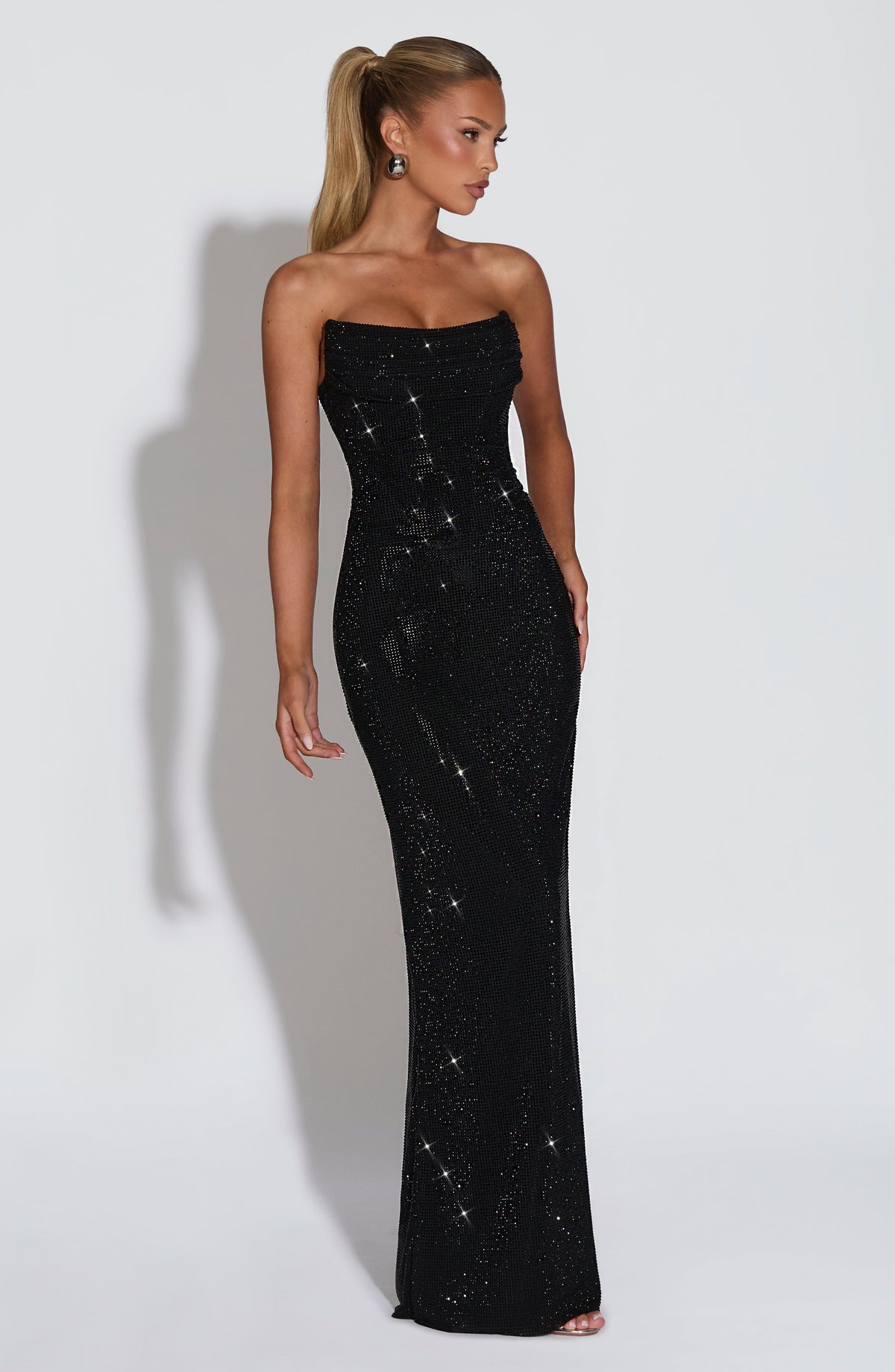 Model wearing the black strapless rhinestone maxi dress, angled three-quarter view showing the fitted bodice and floor-length column skirt. 25
