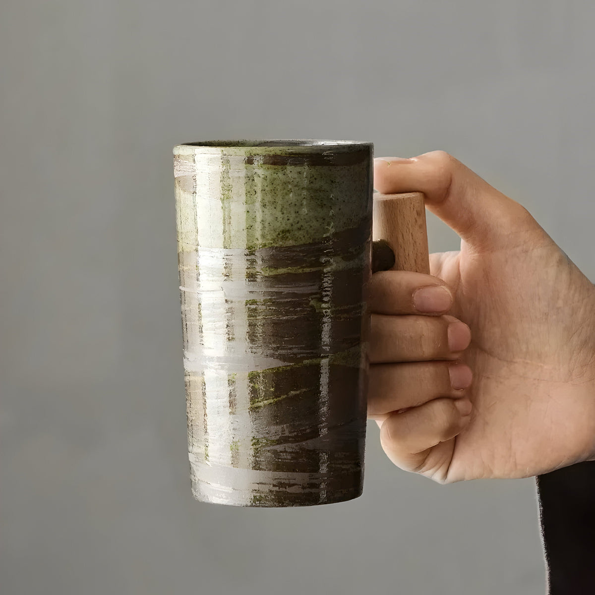 Hand gripping the wooden handle of the green-brown brushed glaze mug, showing its tall size 9