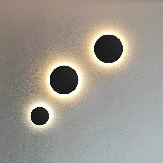 Three black round disc lights in graduated sizes glowing warmly on a pale textured wall. 2