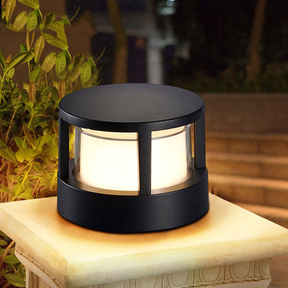 Round black post light with a flat top and glowing white diffuser, standing on a stone pillar beside garden plants at night. 1