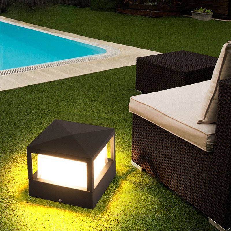 Square black light with a pyramid-style roof standing on a lawn, casting warm glow near a swimming pool and rattan sofa. 4