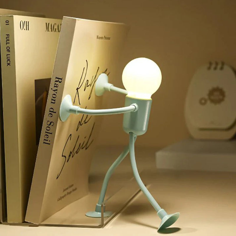 Green figure lamp braces its suction-cup hands against a standing book as if pushing it, lit bulb head glowing warmly. 9