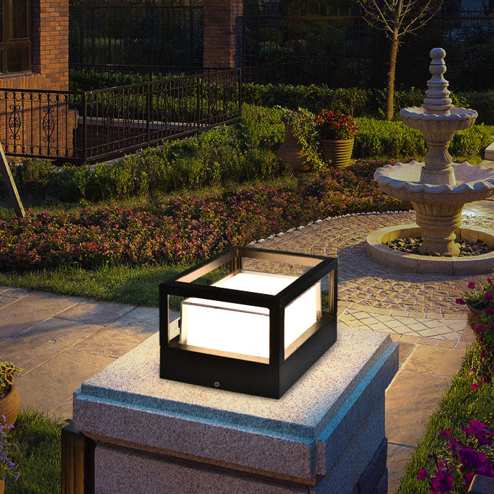 Square open-frame black post light glowing on a granite pillar in a landscaped garden with a stone fountain at dusk. 2
