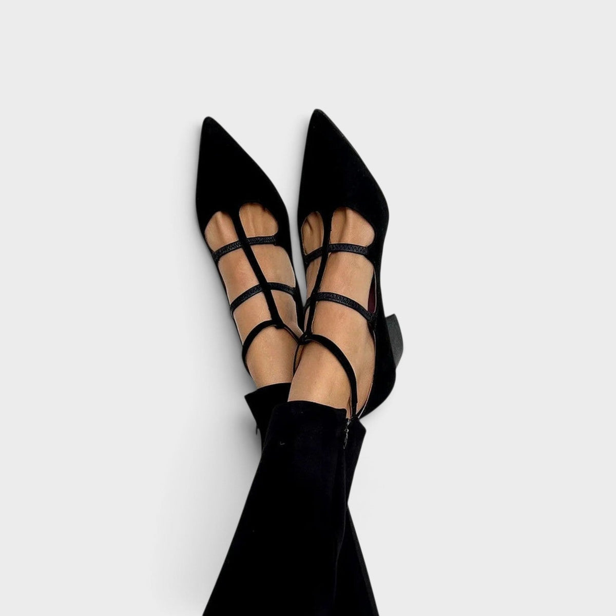 Worn by a model with legs crossed, black pointed-toe heels with a T-bar, two cross straps and an ankle strap, shown with black trousers. 1