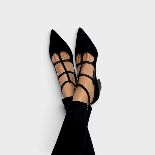 Worn by a model with legs crossed, black pointed-toe heels with a T-bar, two cross straps and an ankle strap, shown with black trousers. 1