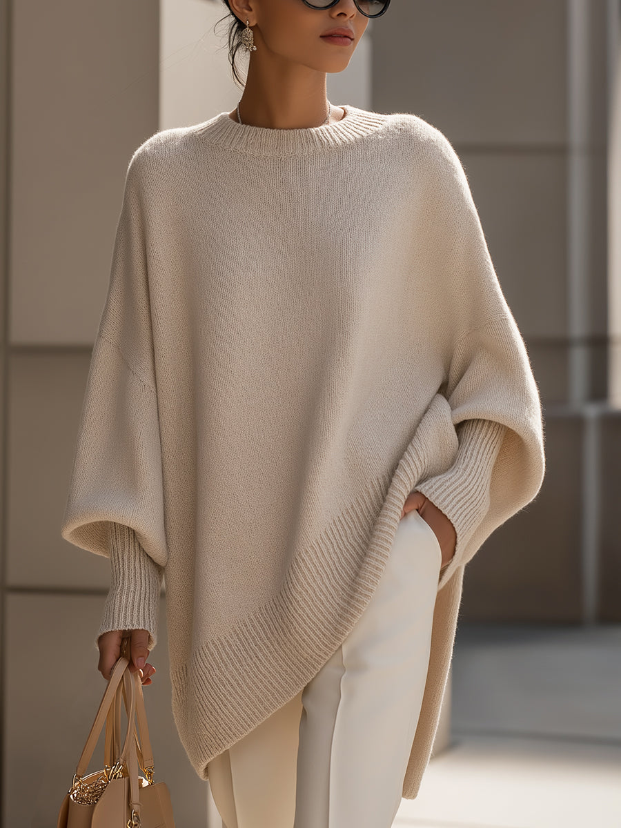 Cream knit poncho in a closer front view, worn with white trousers and one hand in a pocket 2
