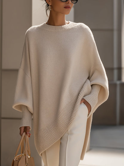 Cream knit poncho in a closer front view, worn with white trousers and one hand in a pocket 2