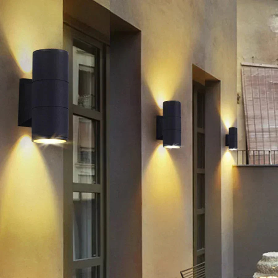 Row of black cylindrical wall lights of differing sizes along a house facade beside windows, casting warm light up and down. 3