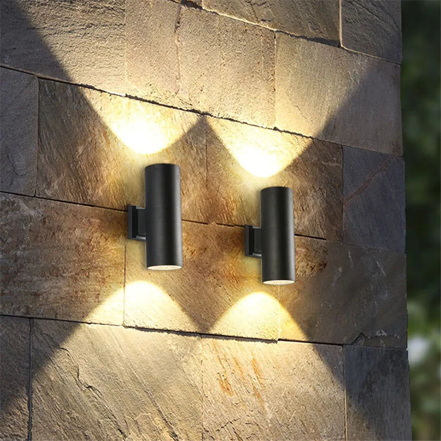 Two black cylindrical wall lights on a rough stone wall, each casting a warm glow upwards and downwards. 2