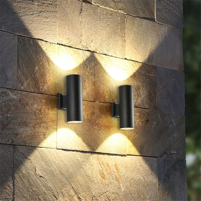 Two black cylindrical wall lights on a rough stone wall, each casting a warm glow upwards and downwards. 2