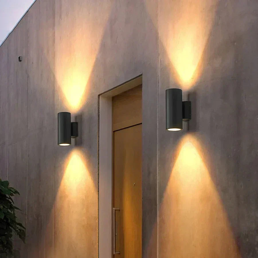 Pair of dark cylindrical wall lights either side of a wooden front door, casting warm cones of light up and down the wall. 6