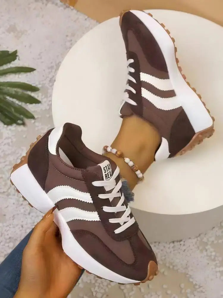 Brown trainers with white stripes, one held in a hand above a white round stand, the other worn 10