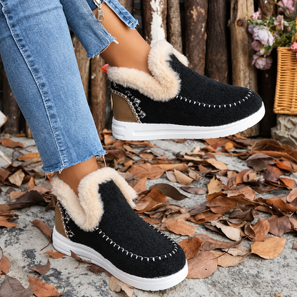 Black textured slip-on trainers with cream plush collars and white contrast stitching, worn with raw-hem jeans among autumn leaves. 1