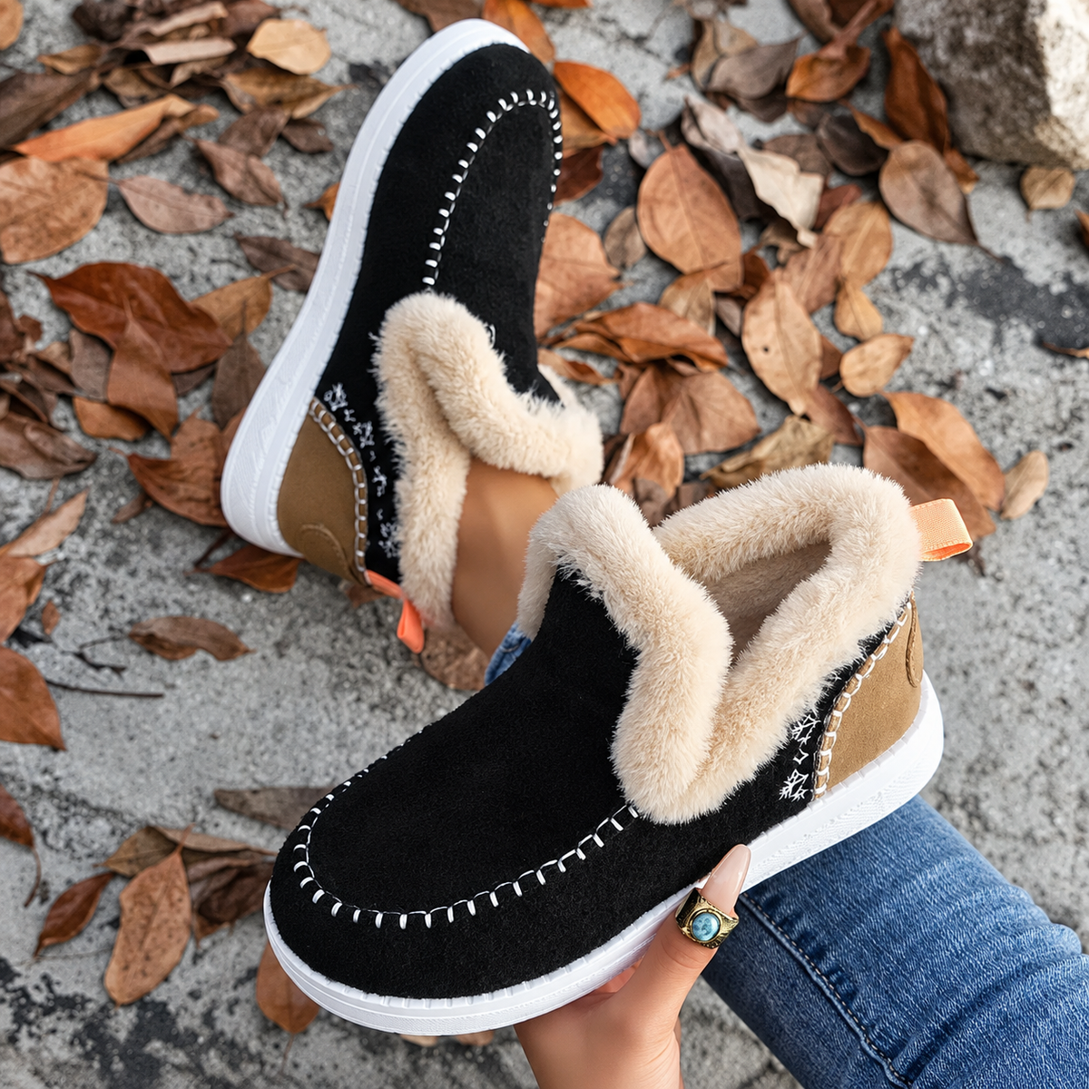 Top-down view of black slip-on trainers with cream plush collars, tan heel panels and snowflake embroidery, held above fallen leaves. 7