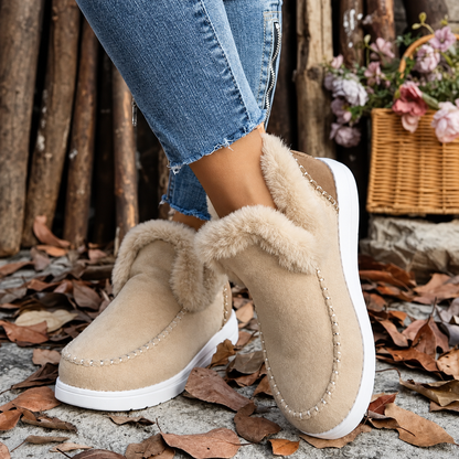 Pair of beige slip-on trainers with fluffy cream collars and white soles, worn with zip-detail jeans beside a flower basket. 9