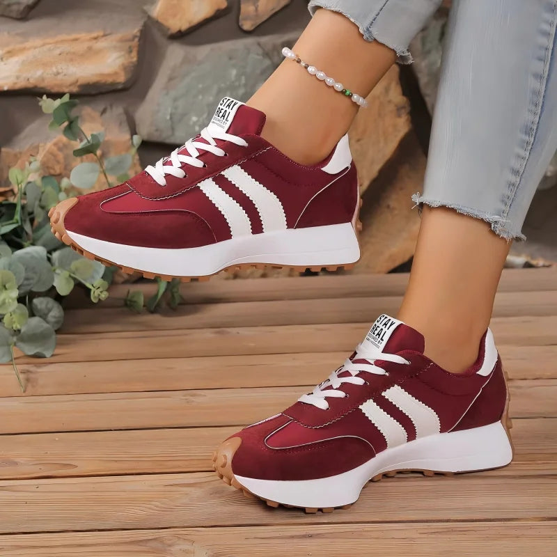 Burgundy trainers with white stripes on a model standing on wooden decking beside green foliage 11
