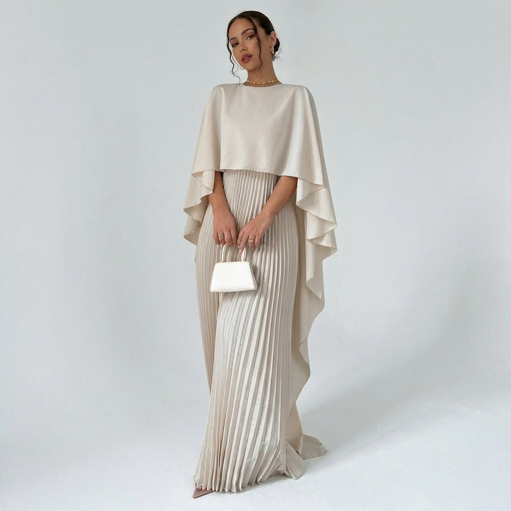 Model in the cream maxi dress with draped cape top and pleated skirt, holding a small white handbag 1