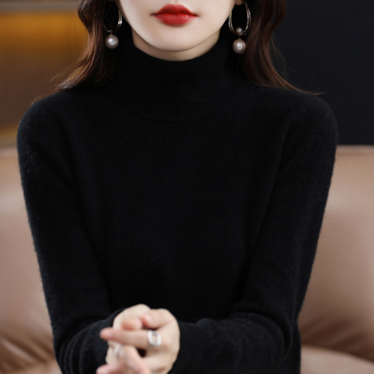 Close-up of a black roll-neck jumper on a model with a short ribbed collar, seated on a tan leather sofa. 2
