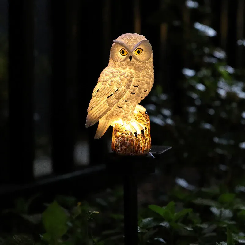 Close view of the white owl light with yellow eyes, lit warmly at night beside dark fencing and foliage. 6