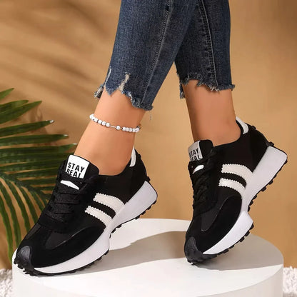 Front-angled view of black trainers with white stripes on a model standing on a white plinth, palm leaf behind 14