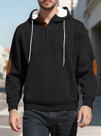 Model walking outdoors in a black diamond-textured hoodie, front view with tan elbow patches and buttons 16