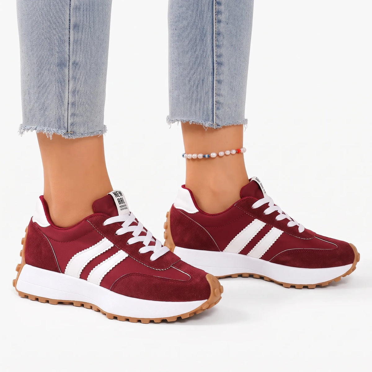 Burgundy trainers with white stripes and a pearl anklet, worn with light jeans on a white background 17