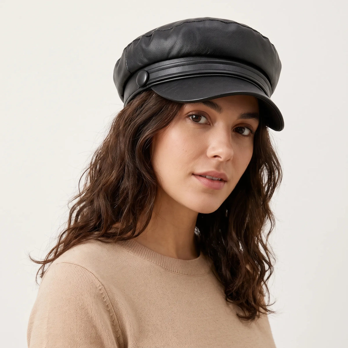 Woman with wavy brown hair wearing the black faux leather baker boy hat with a covered button, turned to the camera 1