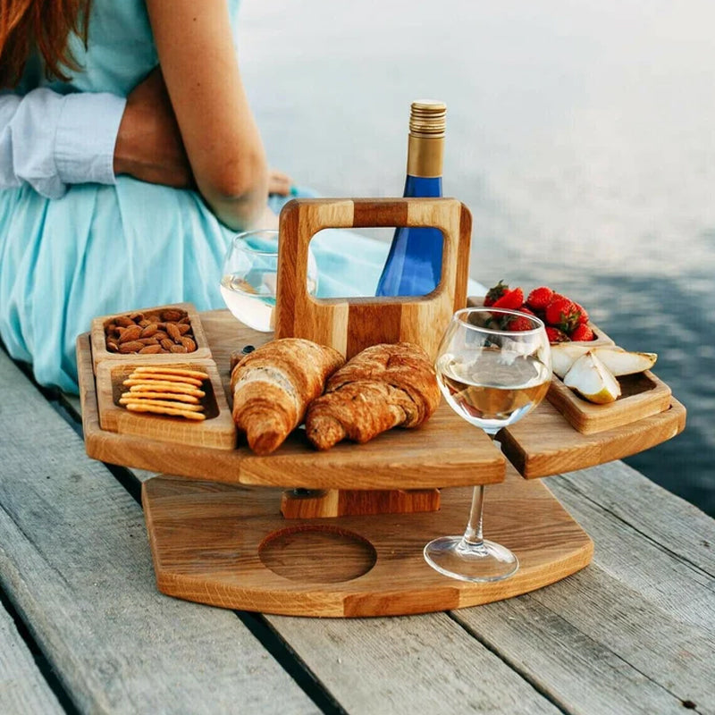 Wooden table with croissants, nuts, fruit, wine and glasses, set on a jetty beside a seated couple 2