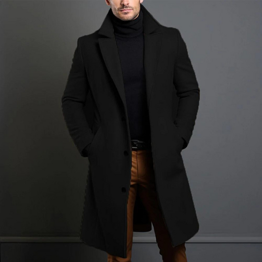 Model wearing the black longline single-breasted coat with notch lapels over a black roll-neck, hands in pockets, cropped at the chin. 1
