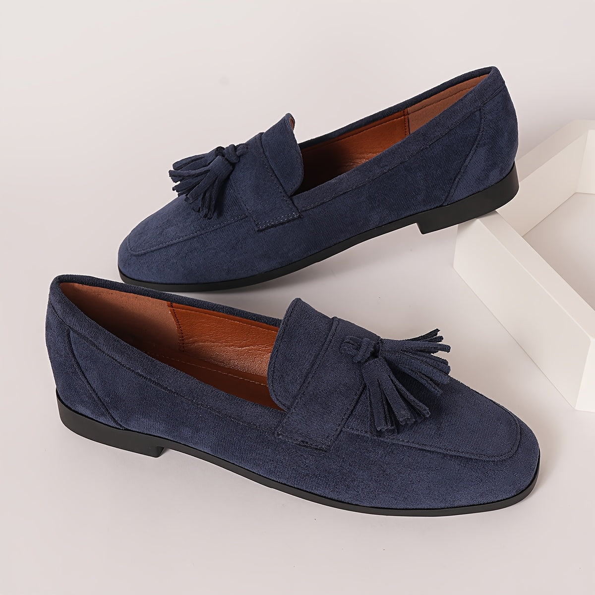 Pair of navy tasselled loafers on a white block, showing tan lining and low black heel 6