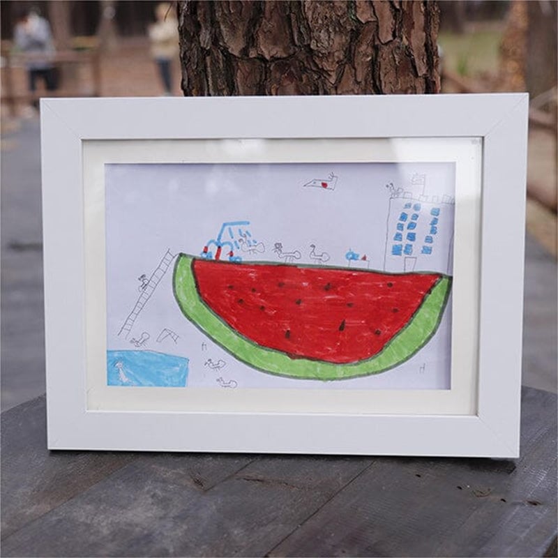 White frame standing on a wooden table outdoors, displaying a child's felt-tip drawing of a giant watermelon slice with tiny figures and a tall building. 21