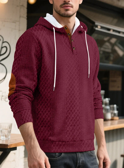 Model in a burgundy diamond-textured hoodie in a cafe setting, three-quarter view with tan elbow patch and buttons 20