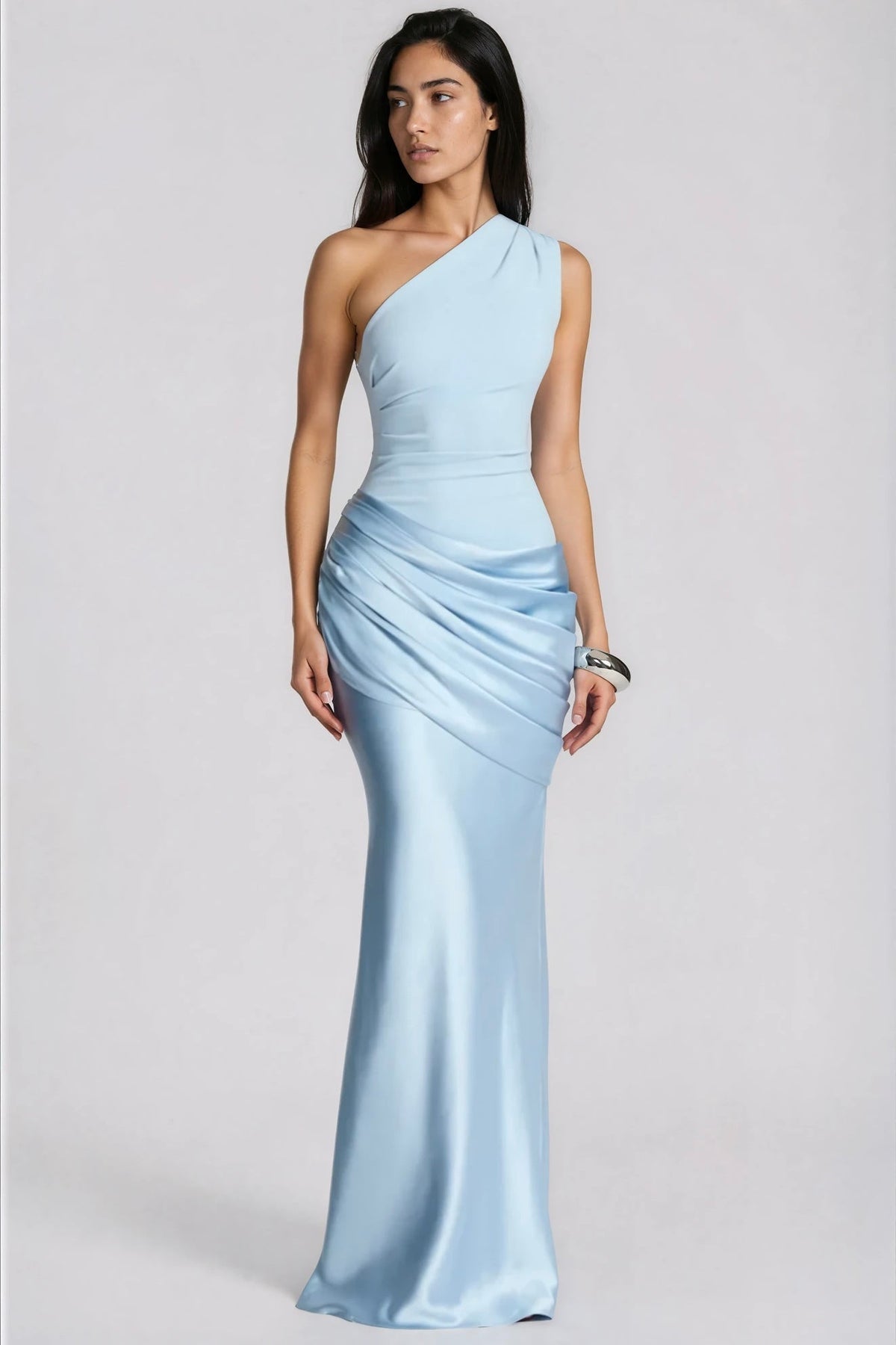 Model in a pale blue one-shoulder maxi dress with draped satin hip panel, looking to the side, wearing a chunky silver cuff. 1