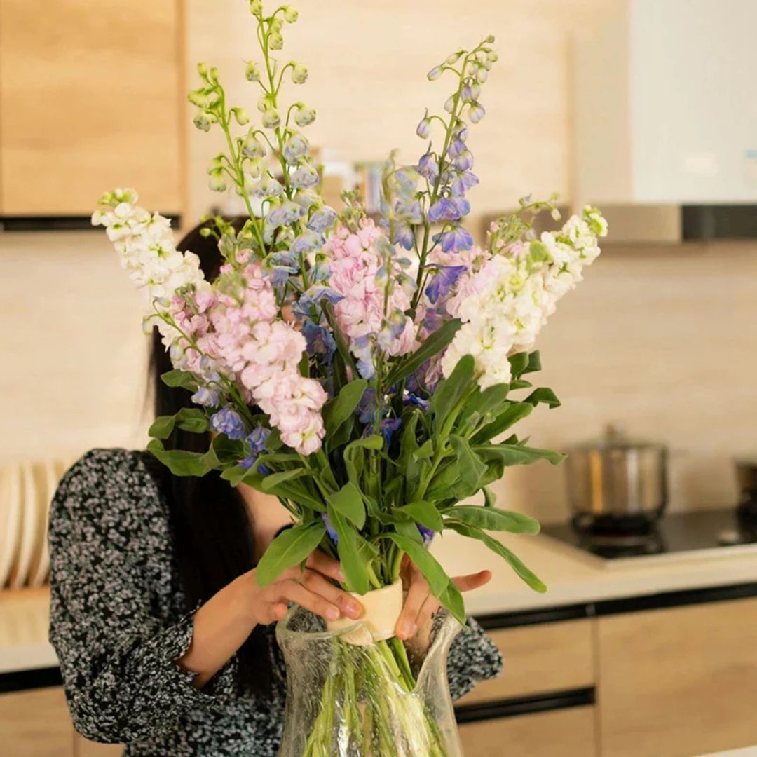 Woman lifts an upright bouquet of pink, white and blue flowers in a glass vase, the cream insert holding the stems together. 2
