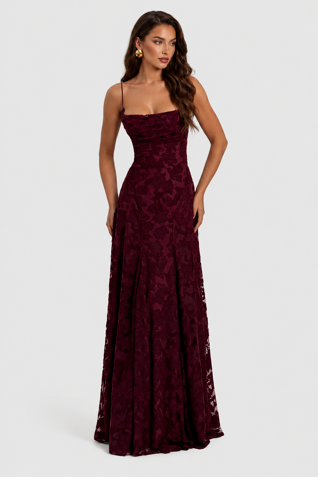 Model in a burgundy raised floral lace gown with draped neckline and gold earrings, hands resting at her sides. 20