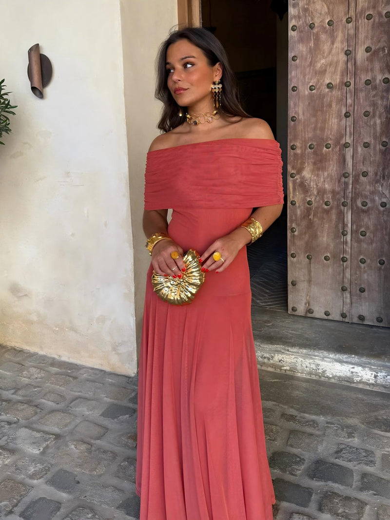 Three-quarter front view of the terracotta off-shoulder dress with gathered neckline, holding a gold shell clutch 7