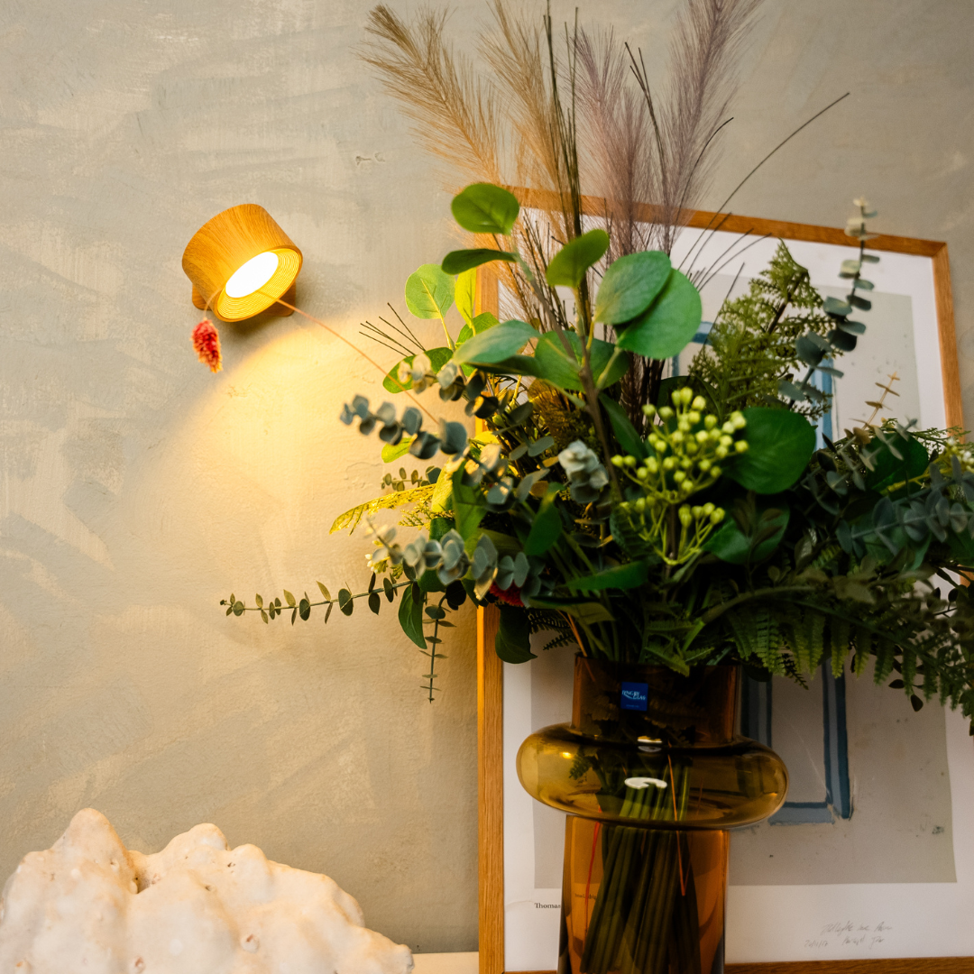 Wood-effect wall light glowing beside a framed print and a vase of eucalyptus, ferns and dried grasses. 6