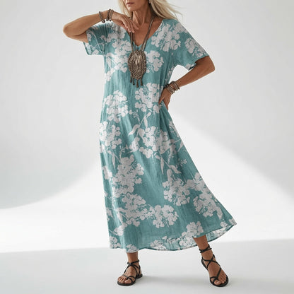 Model in a loose teal V-neck dress with a white blossom print and short sleeves, finished with a chunky pendant necklace and sandals. 4