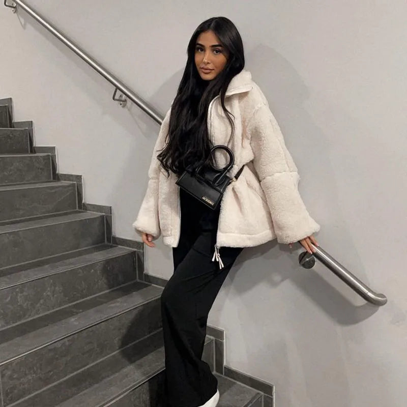 Woman in a cream fleece coat leaning on a stairwell handrail, wearing black trousers and a small black handbag 2