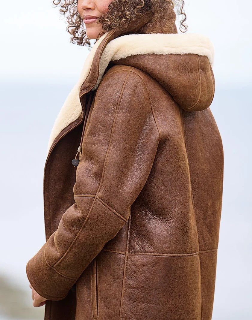 Side view of the coat on a model, showing the lined hood, cream fleece collar, stitched sleeve seams and textured brown outer. 2