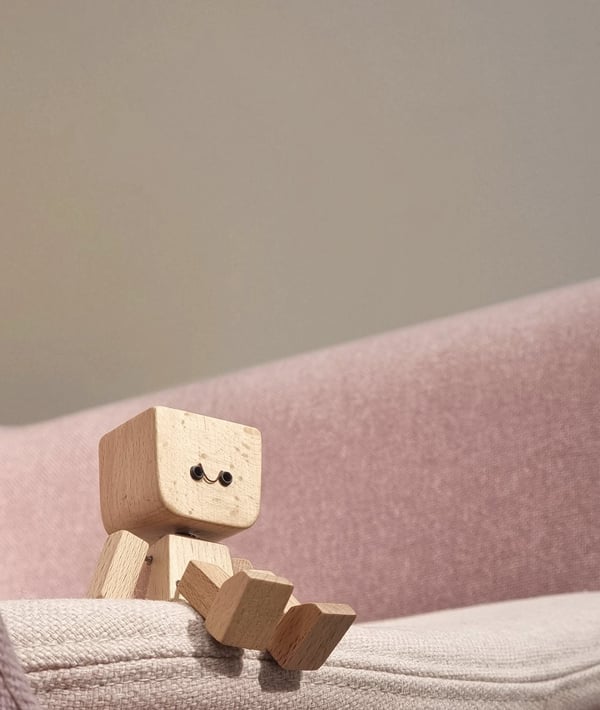 Small wooden figure with a smiling face sitting on the edge of a pink fabric sofa. 4