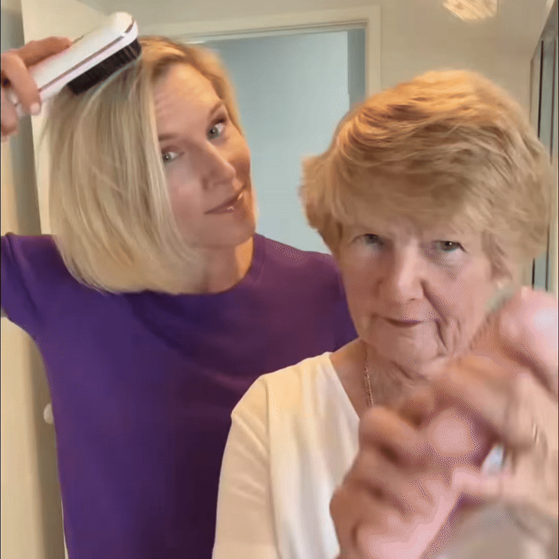 Blonde woman in a purple top using a white brush on her hair while an older woman with short hair stands in front. 9