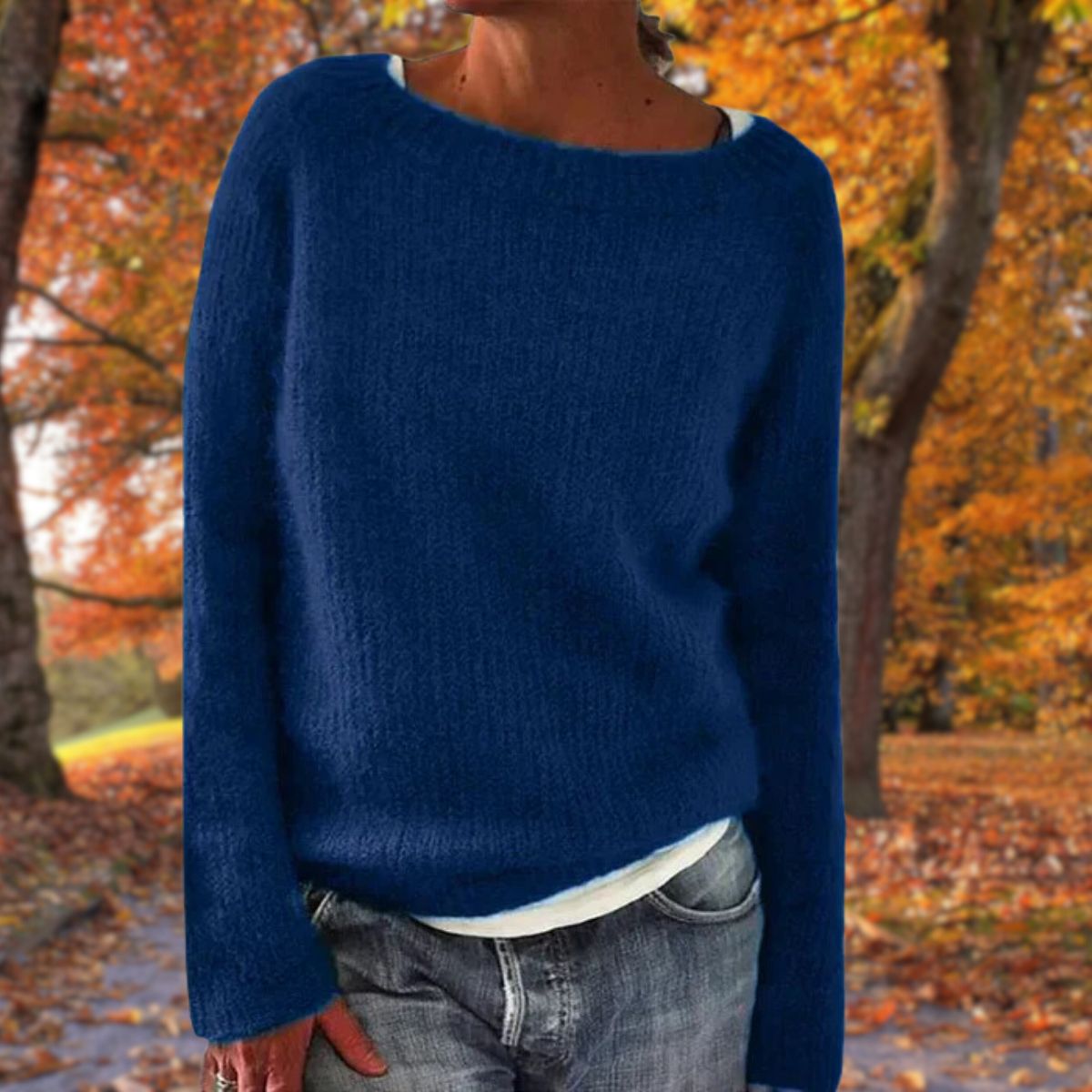 Model in the navy blue version of the soft knit jumper, layered over a white top with jeans among autumn leaves. 3