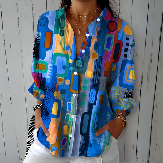 Model wearing the blouse in blue with an abstract geometric print, hands in the pockets of white trousers, layered gold necklaces. 1