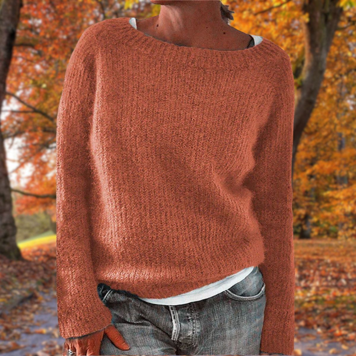 Model in the rust-orange version of the knit jumper, worn over a white top and jeans against autumn foliage. 6