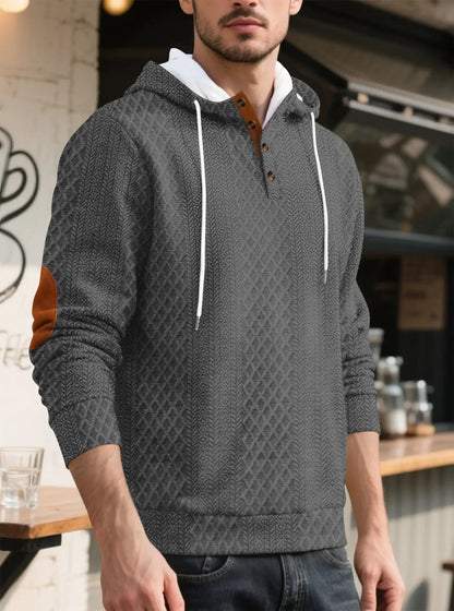 Grey textured hoodie worn by a model in three-quarter view outside a cafe, with tan elbow patch 32