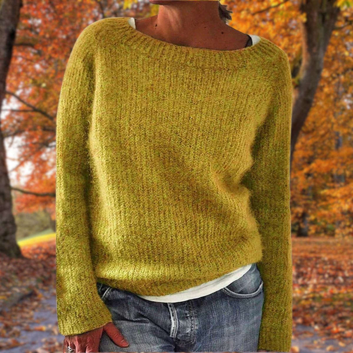 Model in a mustard-yellow knit jumper with a wide neckline, worn over a white top and jeans against autumn trees. 1